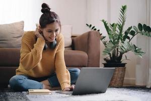 Smiling young woman working on laptop sitting on the floor in front of the sofa wearing headphones at home photo