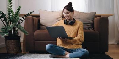 Smiling young woman working on laptop sitting on the floor in front of the sofa wearing headphones at home photo