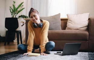 una chica sonriente mirando la cámara junto a la laptop se sienta en el suelo frente al sofá y se pone los auriculares. vacaciones en casa foto