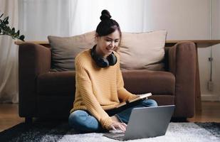 Smiling young woman working on laptop sitting on the floor in front of the sofa wearing headphones at home photo