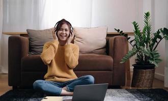 una chica sonriente mirando la cámara al lado del portátil se sienta en el suelo frente al sofá y se pone los auriculares para escuchar música en casa. foto