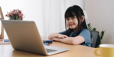 smiling little girl looking at the camera sitting at a table studying at home cute kid doing homework holding a pen taking notes watching webinars photo