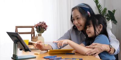Loving senior grandmother hug shoulders of small girl grandchild assist in preparing homework support give advice. Friendly aged woman teacher watching little kid pupil writing maths problem solution