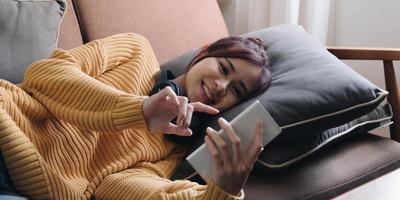Lazy young woman spend weekend at home lying down on couch wearing headphones looking at smart phone screen listening music online. photo