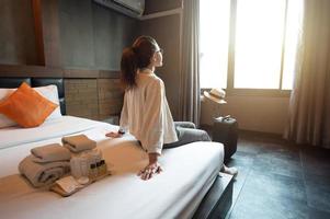 mujer turista sentada en la cama con su equipaje en la habitación del hotel después del check-in y mirando a la hermosa vista exterior de la ventana. conceptual de viaje y alojamiento. foto