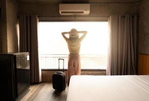 vista trasera de una turista parada cerca de la ventana, mirando a una hermosa vista con su equipaje en la habitación del hotel después del check-in. conceptual de viajes y vacaciones. foto