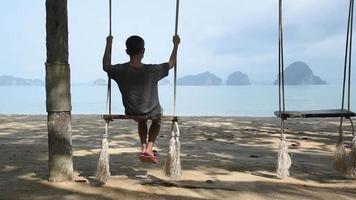 A senior adult Asian wearing face mask sitting on wooden swing and looking at scene of beautiful beach at Krabi province, Thailand video