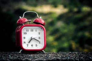 Red retro style alarm clock on a concrete surface with blurry trees on the background. photo