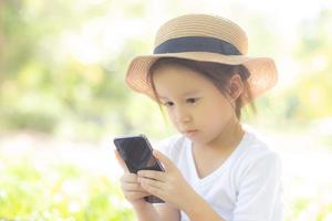 hermoso niño asiático sonriendo usando un teléfono móvil inteligente en el jardín, el niño tiene pasión por jugar juegos digitales en un teléfono inteligente en la red de Internet en el parque con un concepto feliz, de estilo de vida y tecnológico. foto