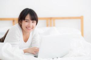 Beautiful portrait asian young woman smile lying on bed using laptop computer with happy at bedroom for leisure and relax, freelance with girl working notebook, communication concept. photo