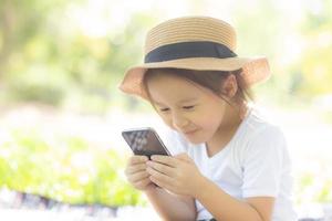 hermoso niño asiático sonriendo usando un teléfono móvil inteligente en el jardín, el niño tiene pasión por jugar juegos digitales en un teléfono inteligente en la red de Internet en el parque con un concepto feliz, de estilo de vida y tecnológico. foto