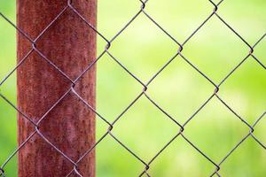A mesh cage in a garden and a rusty pole with green grass as a background. Metal fence with wire mesh. Blurred view of the countryside through a steel iron mesh fence. Abstract background. photo