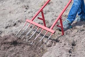 A farmer in jeans digs the ground with a red fork-shaped shovel. A miracle shovel, a handy tool. Manual cultivator. The cultivator is an efficient hand tool for tillage. Loosening the bed. photo