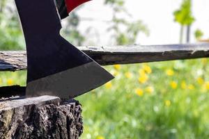 A new modern ax with a red handle sticks out of a wooden stump against a green meadow on a summer sunny day. The ax stuck into the stump. Ax blade in a log.