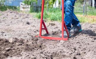 A farmer in jeans digs the ground with a red fork-shaped shovel. A miracle shovel, a handy tool. Manual cultivator. The cultivator is an efficient hand tool for tillage. Loosening the bed. photo