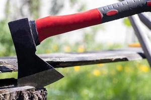 un nuevo hacha moderna con un mango rojo sobresale de un tocón de madera contra un prado verde en un día soleado de verano. el hacha se clavó en el tocón. hoja de hacha en un registro. foto