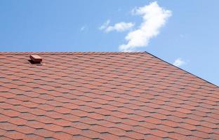 New roof with red shingles against the blue sky. High quality photo. Tiles on the roof of the house. Use to advertise roof fabrication and maintenance. Spotted texture. Affordable roofing. photo