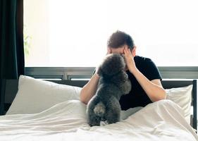 An adorable young black Poodle dog kissing the owner who feel sad and serious on the bed after wake up in the morning with sunshine on messy bed. photo