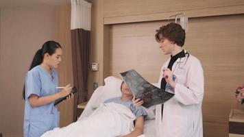 Caucasian female doctor in uniform diagnosis explains x-ray film with Asian radiologist and recovery male patient at inpatient room bed in a hospital ward, medical clinic, cancer examination consult. video