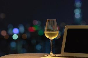 una copa con vino blanco se pone en el sofá con una computadora portátil con una luz colorida de la ciudad.