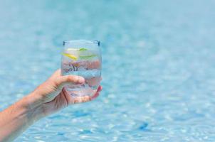 Hand holding a glass of cold drinking water with a piece of lemon on blue color background of swimmin pool. photo