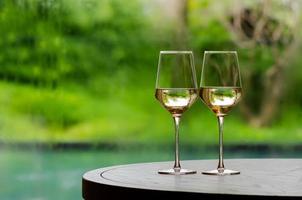 dos copas de vino blanco sobre una mesa de madera para celebrar para una pareja con piscina borrosa y fondo de jardín. foto