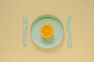 Cutting fresh orange fruit on green plate with knife and fork on yellow background. Minimal creative healthy diet food concept. photo