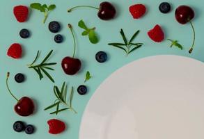 Fresh cherries, blueberries, raspberries, mint and rosemary leaf on top view with white plate and pastel blue color background for healthy food concept. photo