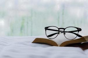 enfoque selectivo en espectáculos y libros puestos en la cama por la mañana con gotas de lluvia en la ventana. quédate en casa y relaja el concepto. foto