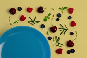 cerezas frescas, arándanos, frambuesas, hojas de menta y romero en la vista superior con plato azul y fondo de color amarillo pastel para un concepto de comida saludable. foto