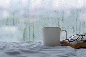 A cup of coffee with book and spectacles on bed in morning with rain drop on window. Stay home and relaxing concept. photo