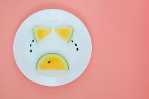 Crying face that eyes and mouth made from yellow watermelon with tear made from its seeds put on a white plate with pink background. photo