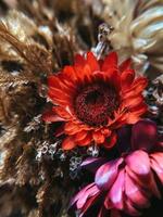 Red beautiful artificial flower on textured blurred background with dried plants, vertical photo. Decorative design interior element, closeup, wallpaper photo