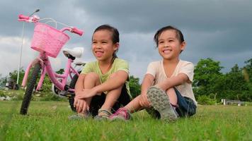 feliz menina bonitinha e sua irmã sentada no gramado perto das bicicletas no parque. crianças descansando depois de andar de bicicleta. atividades saudáveis de verão para crianças
