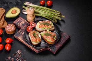 Toast sandwich with butter, avocado and salmon, decorated with arugula and sesame seeds, on a black stone background