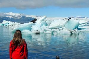 Girl standing on the edge of the Jokulsarlon lake, facing bright clear blue icebergs floating in the blue cold water, in Iceland 2 photo