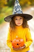 A girl in a witch's hat, in a yellow sweater and with a pumpkin with carved eyes and a mouth Jack o Lantern in a yellow autumn forest. Halloween Holiday photo