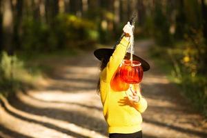 A girl in a witch's hat, in a yellow sweater and with a pumpkin a lantern and a bucket for sweets in autumn forest. Halloween Holiday. Portrait, close-up photo