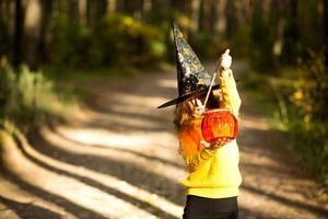 A girl in a witch's hat, in a yellow sweater and with a pumpkin a lantern and a bucket for sweets in autumn forest. Halloween Holiday. Portrait, close-up photo