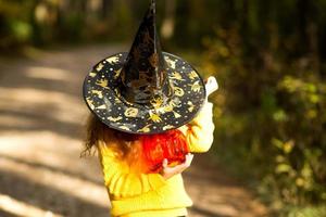 una chica con un sombrero de bruja, un suéter amarillo y una calabaza, una linterna y un balde para dulces en el bosque de otoño. vacaciones de halloween retrato, primer plano foto