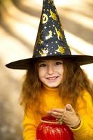 A girl in a witch's hat, in a yellow sweater and with a pumpkin a lantern and a bucket for sweets in autumn forest. Halloween Holiday. Portrait, close-up photo