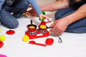 Dad and daughter play with sliced plastic vegetables and fruits with Velcro, cooks food on a toy stove in a bowl. Children's cooking, a girl learns to cook. White background, close-up. Father's Day photo