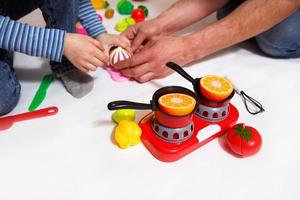 Dad and daughter play with sliced plastic vegetables and fruits with Velcro, cooks food on a toy stove in a bowl. Children's cooking, a girl learns to cook. White background, close-up. Father's Day photo