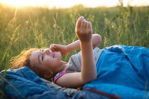 la niña no está satisfecha con las picaduras de mosquitos, el niño duerme en un saco de dormir sobre la hierba en un viaje de campamento. recreación al aire libre ecológica, horario de verano. alteración del sueño, repelente. foto