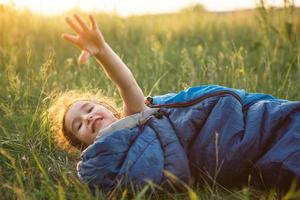 A child sleeps in a sleeping bag on the grass in a camping trip - eco-friendly outdoor recreation, healthy lifestyle, summer time. Sweet and peaceful sleep. Mosquito bites, repellent. photo