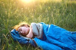 A child sleeps in a sleeping bag on the grass in a camping trip - eco-friendly outdoor recreation, healthy lifestyle, summer time. Sweet and peaceful sleep. Mosquito bites, repellent. photo