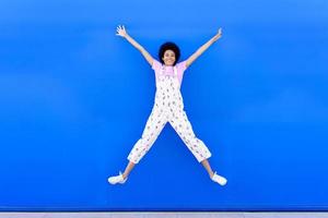 mujer afroamericana saltando con los brazos levantados contra una pared urbana azul. foto