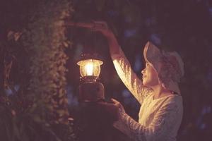 asian girl in a white dress Holding an ancient lantern in her hand, she looked forward in the dark forest at night.soft focus. photo