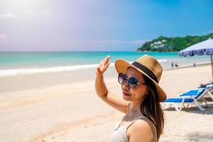 Young woman traveler wearing sunglasses covering face by hand to protect UV rays from the sun at tropical sandy beach on sunny day, Skin care and eyes protect concept photo