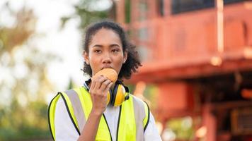 smile African american woman foreman worker  or woman maintenance engineer in reflective vest safety jacket sits down on old truck for relaxing, eats bread snacks and water during brunch break photo